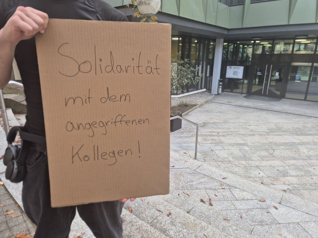 Eine Person hält eine Papptafel mit dem Text "Solidarität mitdem angegriffenen Kollegen" in die Kamera. Im Hintergrund ist das Gebäude des Landgerichts in der Olgastraße 2 zu sehen, in dem der Prozess stattfindet